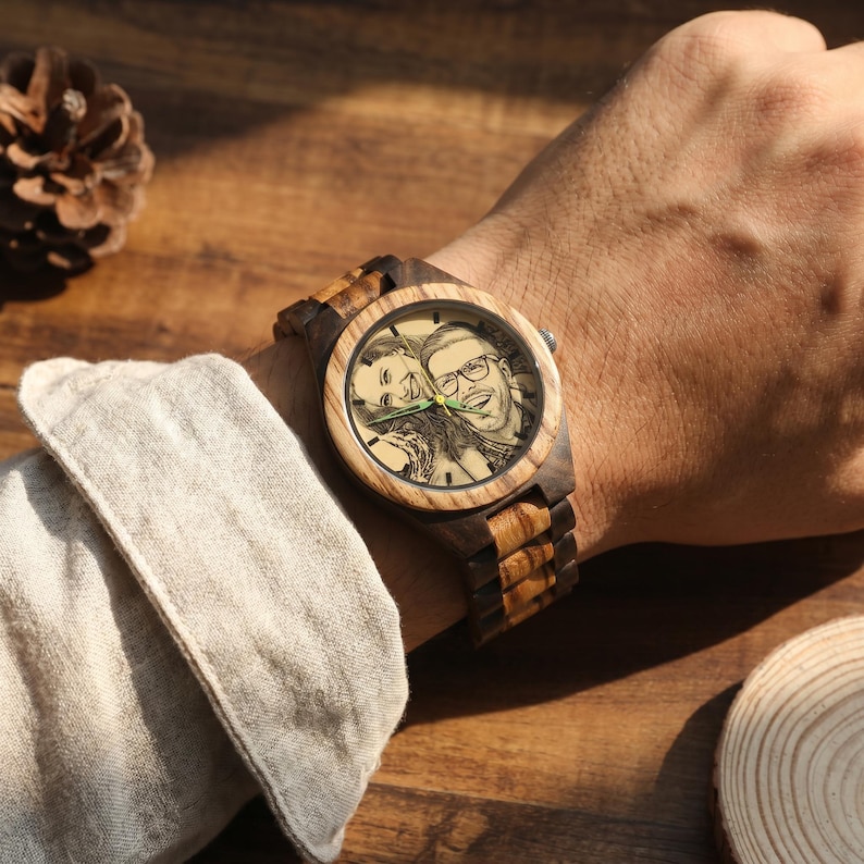 May include: A wristwatch with a wooden case and link band featuring a light and dark brown striped grain pattern. The watch face displays a custom engraved black and white portrait of two people, with thin green hour, minute, and second hands.
