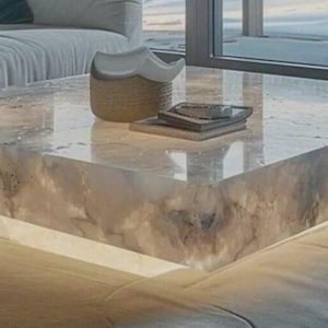 May include: A modern, square coffee table with a translucent, marbled surface. The table has a built-in, glowing base, and is topped with a decorative basket and a stack of books. The table is approximately 48 inches square.