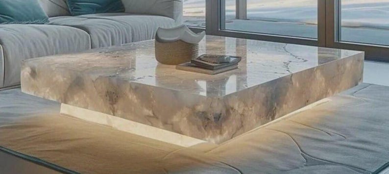 May include: A modern, square coffee table with a translucent, marbled surface. The table has a built-in, glowing base, and is topped with a decorative basket and a stack of books. The table is approximately 48 inches square.