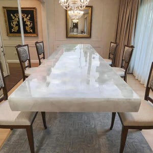 May include: A long rectangular dining table with a polished white translucent stone surface featuring natural veining. The table is surrounded by six chairs with dark wooden frames, light beige upholstered seats, and circular backrest patterns.