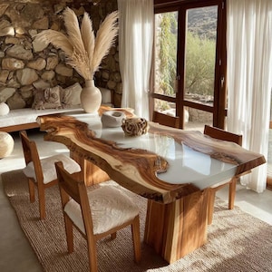 Op de afbeelding: Een houten eettafel met een natuurlijke rand en een witte harsinleg. De tafel wordt ondersteund door twee houten sokkels. Twee houten stoelen met witte, donzige zitkussens staan op een geweven tapijt.