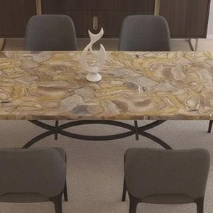 Op de afbeelding: Een rechthoekige eettafel met een patroon van lichtbruin en beige steen. De tafel heeft een donker metalen onderstel en is omringd door zes grijze gestoffeerde stoelen. Een witte decoratieve sculptuur staat op de tafel.