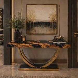 Puede incluir: Una mesa consola con un tablero rectangular de mármol negro y ámbar, sostenida por dos patas de metal dorado curvadas sobre una base negra. La mesa sostiene un jarrón oscuro acanalado con follaje verde, una vela y tres libros con un cuenco dorado.