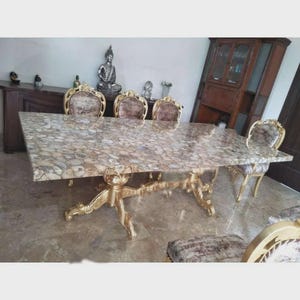 May include: A large, rectangular dining table with a patterned stone top and ornate gold-colored legs. Six matching chairs with gold frames and patterned upholstery surround the table. A Buddha statue and other decorative items are visible in the background.