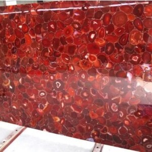 May include: A large, rectangular slab of polished red agate stone. The surface is a mosaic of rounded, reddish-brown stones with lighter, circular patterns. The overall effect is a vibrant, textured surface, suitable for countertops or decorative panels.