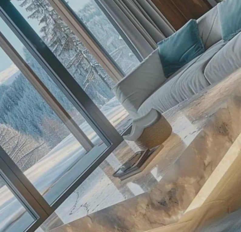 May include: A modern living room with a large window overlooking a snowy mountain landscape. A white sofa with a teal pillow sits near the window. A light-colored, marble-like coffee table holds a woven basket and a book.