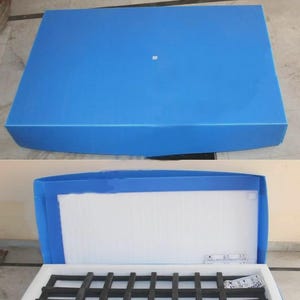 May include: Blue packing box with the text "PACKING BOX" in bold white letters. The box is open, revealing a white interior with a black grid-like structure. The box is designed for shipping and storage.