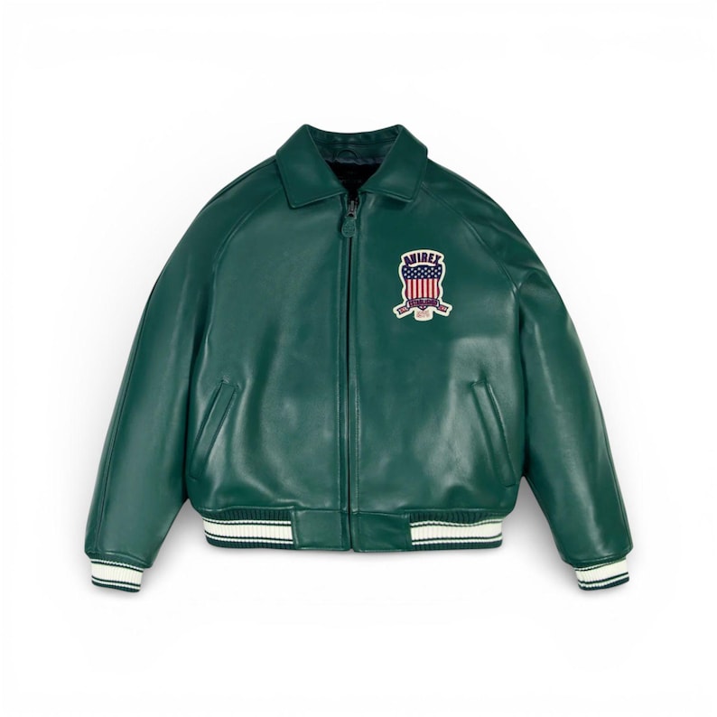 Puede incluir: Chaqueta bomber de cuero verde oscuro con cremallera y cuello. La chaqueta presenta un parche en el pecho con la palabra "AVIREX" y un dise&ntilde;o que incorpora la bandera estadounidense. Los pu&ntilde;os y el dobladillo tienen rayas blancas y azul marino.