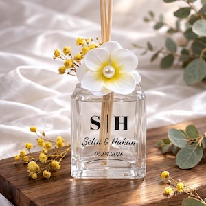 May include: A clear glass diffuser bottle with the initials "S | H" and the names "Selin & Hakan" and the date "05.04.2026" printed on the front. The bottle is decorated with a white flower with a pearl centre and yellow flowers. Wooden reeds are inserted into the bottle.