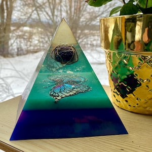 May include: A pyramid-shaped resin art piece featuring a butterfly and a stone. The pyramid is layered with teal, blue, and purple hues. A gold-coloured pot with a plant is visible in the background.