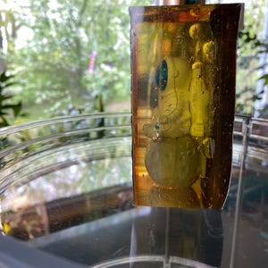 K&ouml;nnte beinhalten: Ein klarer, rechteckiger Harzblock, der eine kleine Astronautenfigur umschlie&szlig;t. Der Astronaut tr&auml;gt einen wei&szlig;en Raumanzug mit blauem Visier und steht auf einem gelben Planeten. Das Harz hat einen gelb-braunen Farbton.