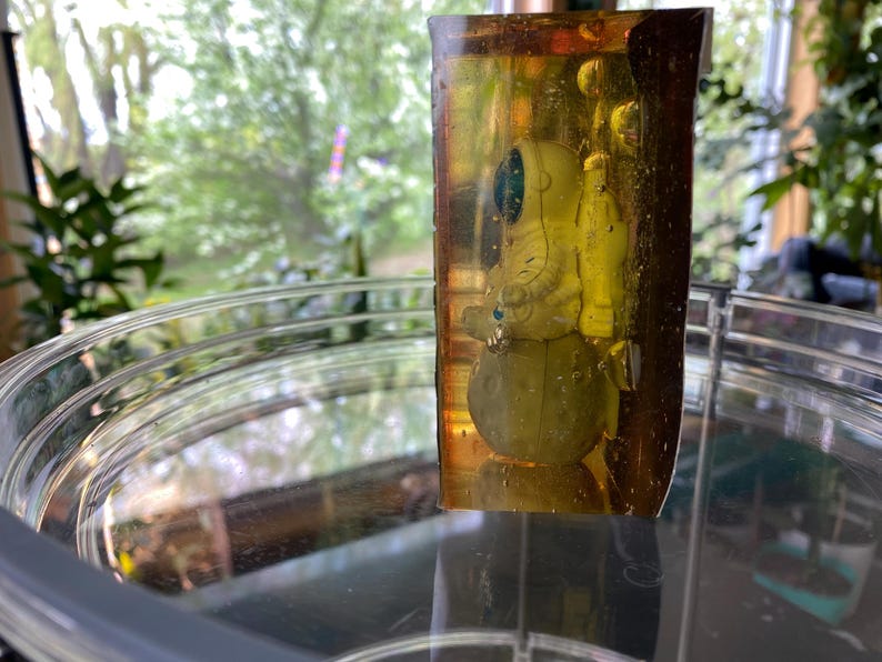 K&ouml;nnte beinhalten: Ein klarer, rechteckiger Harzblock, der eine kleine Astronautenfigur umschlie&szlig;t. Der Astronaut tr&auml;gt einen wei&szlig;en Raumanzug mit blauem Visier und steht auf einem gelben Planeten. Das Harz hat einen gelb-braunen Farbton.