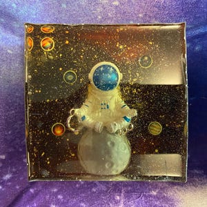 K&ouml;nnte beinhalten: Ein quadratisches Kunstwerk aus Harz mit einem Astronauten, der auf einem Mond meditiert. Der Hintergrund ist eine dunkle Weltraumszene mit Sternen und Planeten. Der Astronaut tr&auml;gt einen wei&szlig;en Raumanzug mit blauen Akzenten.