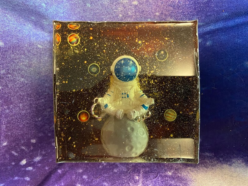 K&ouml;nnte beinhalten: Ein quadratisches Kunstwerk aus Harz mit einem Astronauten, der auf einem Mond meditiert. Der Hintergrund ist eine dunkle Weltraumszene mit Sternen und Planeten. Der Astronaut tr&auml;gt einen wei&szlig;en Raumanzug mit blauen Akzenten.