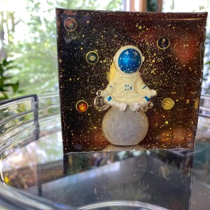 K&ouml;nnte beinhalten: Ein Kunstwerk aus Harz mit einer Astronautenfigur, die auf einem grauen Mond vor einem dunklen, sternen&uuml;bers&auml;ten Hintergrund sitzt. Der Astronaut ist wei&szlig; mit einem blauen Helm. Der Hintergrund hat goldene und gelbe Akzente mit Planeten.