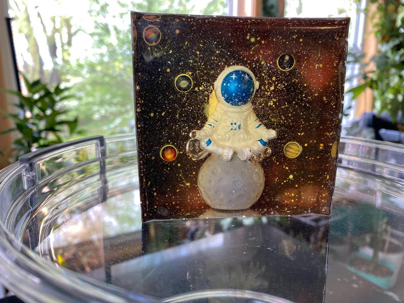 K&ouml;nnte beinhalten: Ein Kunstwerk aus Harz mit einer Astronautenfigur, die auf einem grauen Mond vor einem dunklen, sternen&uuml;bers&auml;ten Hintergrund sitzt. Der Astronaut ist wei&szlig; mit einem blauen Helm. Der Hintergrund hat goldene und gelbe Akzente mit Planeten.