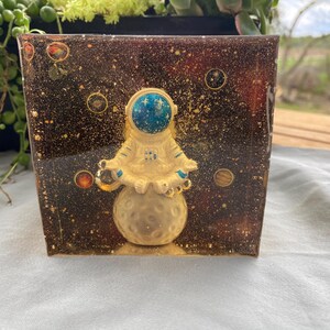 K&ouml;nnte beinhalten: Ein quadratischer Harz-Blumentopf mit Weltraumthema. Der Topf zeigt eine goldene Astronautenfigur, die auf einem Mond sitzt, vor einem dunkelbraunen Hintergrund mit Goldflocken und Planeten. Der Helm des Astronauten ist blau.