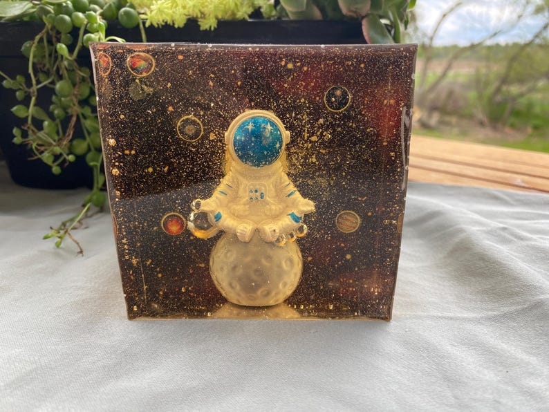 K&ouml;nnte beinhalten: Ein quadratischer Harz-Blumentopf mit Weltraumthema. Der Topf zeigt eine goldene Astronautenfigur, die auf einem Mond sitzt, vor einem dunkelbraunen Hintergrund mit Goldflocken und Planeten. Der Helm des Astronauten ist blau.