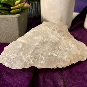 Puede incluir: Un gran cristal de cuarzo translúcido con una forma rugosa e irregular. El cristal es de color blanco lechoso pálido y parece sin pulir. El cristal se exhibe sobre una superficie púrpura, con una planta suculenta y otros elementos decorativos en el fondo.