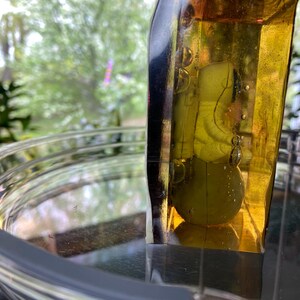 K&ouml;nnte beinhalten: Eine klare Glasschale mit grauem Rand und einem gelb get&ouml;nten Glasobjekt. Das gelbe Glasobjekt hat eine abgerundete Form und enth&auml;lt ein gr&uuml;nes Objekt. Der Hintergrund zeigt eine verschwommene Ansicht von gr&uuml;nem Laub.