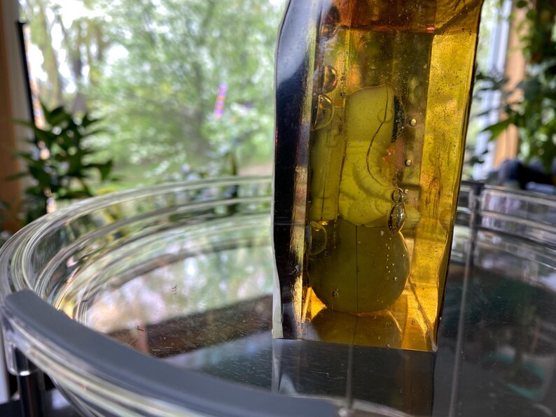 K&ouml;nnte beinhalten: Eine klare Glasschale mit grauem Rand und einem gelb get&ouml;nten Glasobjekt. Das gelbe Glasobjekt hat eine abgerundete Form und enth&auml;lt ein gr&uuml;nes Objekt. Der Hintergrund zeigt eine verschwommene Ansicht von gr&uuml;nem Laub.
