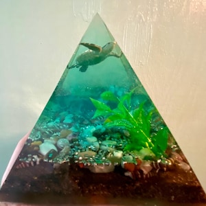 May include: A clear, pyramid-shaped resin sculpture featuring a miniature sea turtle swimming in turquoise water. The base of the pyramid has small pebbles and green plant accents, creating an underwater scene.