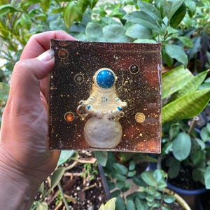 K&ouml;nnte beinhalten: Ein quadratisches Kunstwerk aus Harz mit einem Astronauten in einem Raumanzug, der auf einem Mond steht. Der Hintergrund ist dunkelbraun mit Goldflocken und Planeten. Der Helm des Astronauten ist blau.