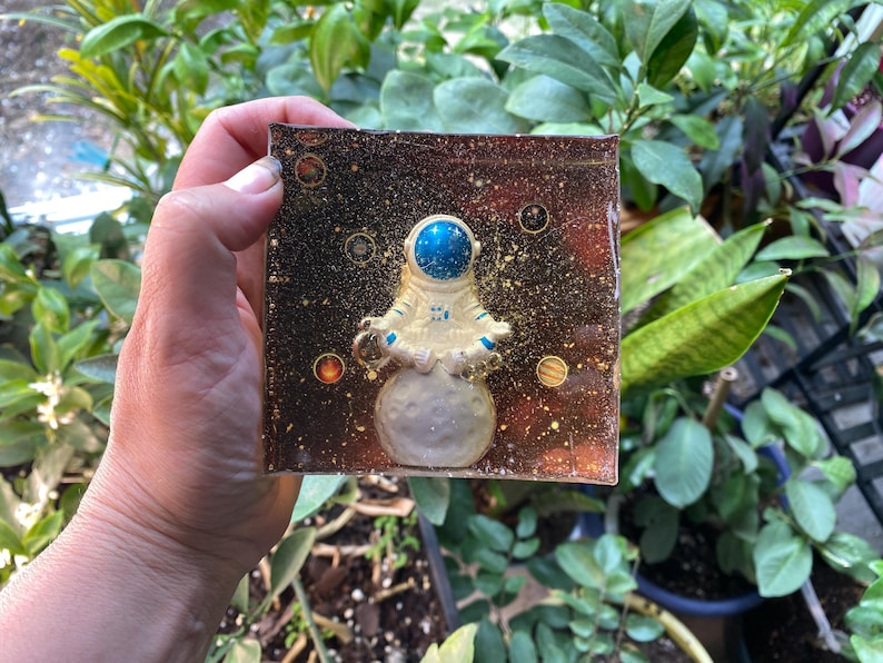 K&ouml;nnte beinhalten: Ein quadratisches Kunstwerk aus Harz mit einem Astronauten in einem Raumanzug, der auf einem Mond steht. Der Hintergrund ist dunkelbraun mit Goldflocken und Planeten. Der Helm des Astronauten ist blau.