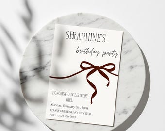 Puede incluir: Una tarjeta de invitación blanca con el texto "SERAPHINE'S birthday party" y "HONORING OUR BIRTHDAY GIRL!" en fuente marrón oscuro. La tarjeta está adornada con un diseño de cinta marrón oscuro y detalles del evento. La tarjeta descansa sobre un plato de mármol.