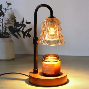 Puede incluir: Una lámpara calentadora de velas con soporte de metal negro, base redonda de madera y pantalla de vidrio ámbar con forma de flor. Una vela en frasco de vidrio con la etiqueta Aroma Sensation se asienta sobre la base, iluminada por la luz.