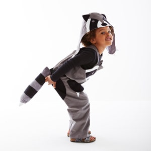 May include: A child wearing a gray and black raccoon costume with a white face and a black tail. The costume has a hood with ears and a black and white face.