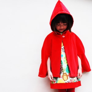 May include: A young girl wearing a red hooded cape with a white trim. She is wearing a colorful dress with a pattern of animals and trees. The girl is looking down at the dress.