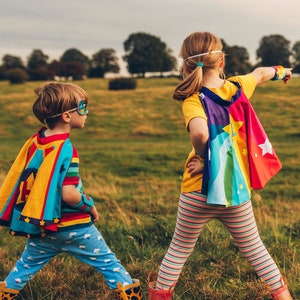May include: Two children wearing colorful capes and superhero masks stand in a grassy field. The child on the left is wearing a blue and yellow cape with stars and a blue mask. The child on the right is wearing a rainbow cape with stars and a white headband.