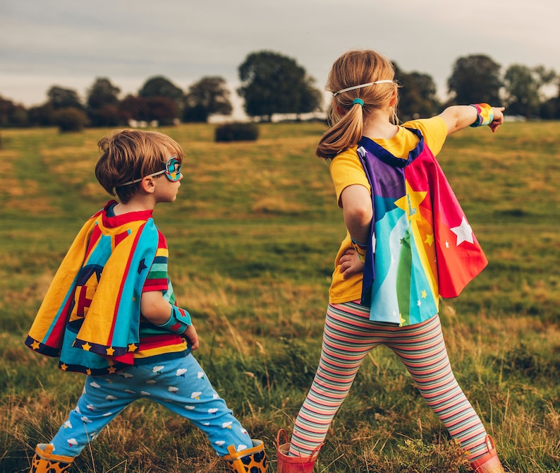 May include: Two children wearing colorful capes and superhero masks stand in a grassy field. The child on the left is wearing a blue and yellow cape with stars and a blue mask. The child on the right is wearing a rainbow cape with stars and a white headband.