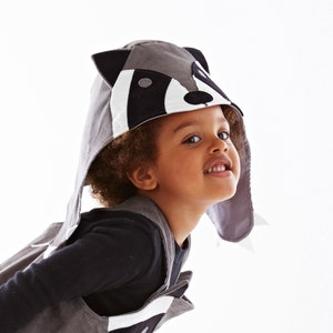 May include: A child wearing a gray raccoon costume with black and white facial features. The costume has a hood and long ears. The child is smiling and looking at the camera.