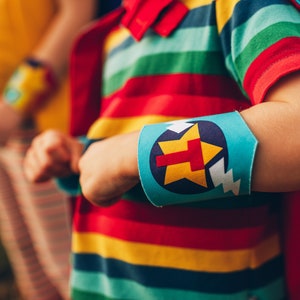 May include: A child wearing a red, yellow, green, and blue striped shirt with a blue and white arm band. The arm band has a yellow star with a red letter T in the center.