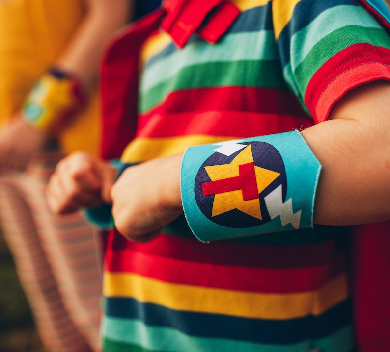 May include: A child wearing a red, yellow, green, and blue striped shirt with a blue and white arm band. The arm band has a yellow star with a red letter T in the center.