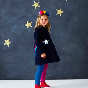May include: A young person wearing a navy blue coat with a white star and a rainbow stripe design. They are also wearing blue and pink leggings, red high-top sneakers, and a colorful pom-pom headband. Gold star decorations are on the dark blue background.