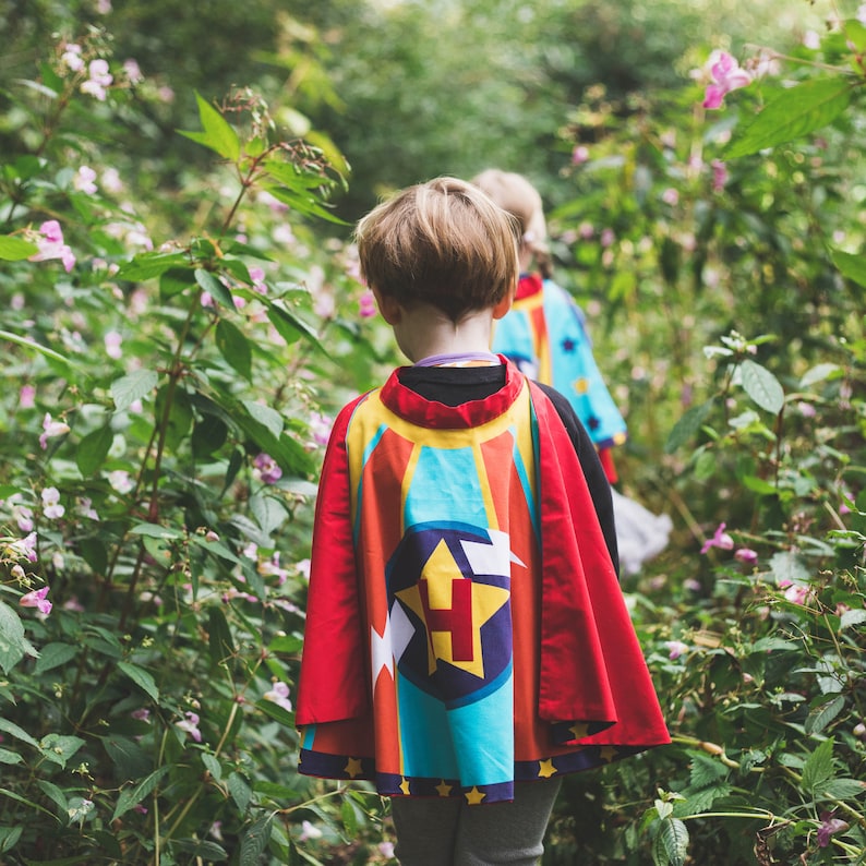 May include: Two children wearing colorful capes with a star and letter H design, walking through a green forest with pink flowers.