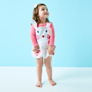 May include: A little girl wearing a white bunny-shaped romper with pink accents and a pink long-sleeved shirt. She is looking up and smiling.
