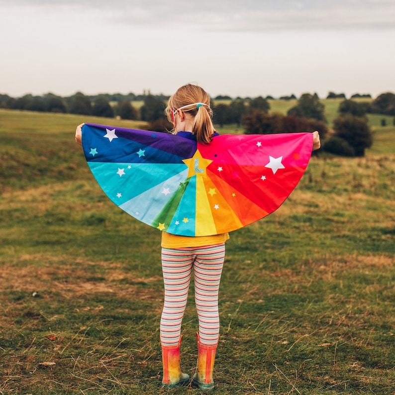 May include: A child wearing a rainbow cape with stars and a letter L on it. The cape is made of fabric and has a rainbow pattern. The child is standing in a field of grass.
