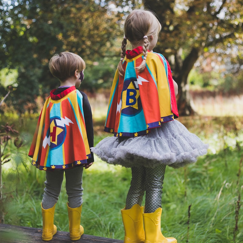 May include: Two children wearing colorful superhero capes with lightning bolts and stars. The capes have a large circle with a letter in the center. The child on the left is wearing a cape with the letter "H" and the child on the right is wearing a cape with the letter "B".