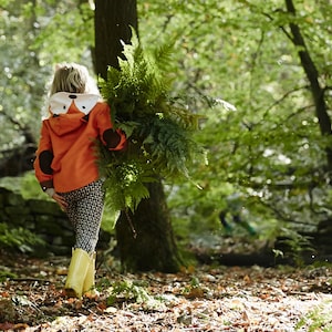 May include: A young person wearing a bright orange hooded jacket with a fox face design walks through a forest carrying a large bunch of ferns. A white dog with a blue and white patterned collar walks beside them.