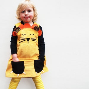 May include: A young child wearing a yellow corduroy dress with a lion face applique. The dress has black paw pockets with white claws. The child is wearing yellow and white striped tights.