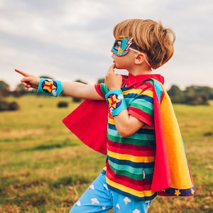 May include: A young child dressed as a superhero wearing a red and yellow cape, a blue mask with a lightning bolt design, and blue and yellow arm bands with a star design. The child is pointing to the right and looking off to the side.