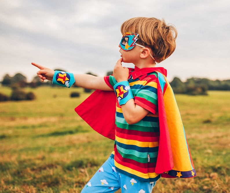 May include: A young child dressed as a superhero wearing a red and yellow cape, a blue mask with a lightning bolt design, and blue and yellow arm bands with a star design. The child is pointing to the right and looking off to the side.