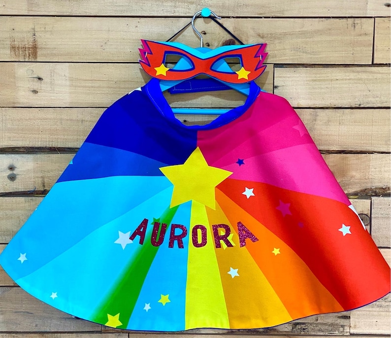 May include: A rainbow cape with a star and the name "AURORA" in pink glitter. The cape is on a hanger with a red and blue superhero mask.