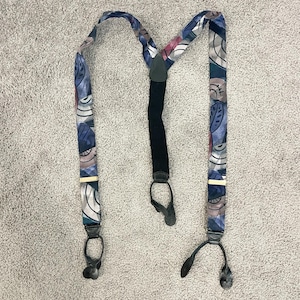 May include: A pair of patterned suspenders with a black elastic back strap and leather details. The suspenders feature a repeating design of blue, gray, and pink abstract shapes. The suspenders have metal clasps for attachment.