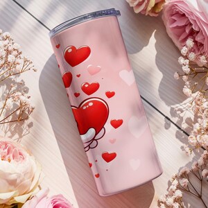 Op de afbeelding: Een roze ge&iuml;soleerde beker versierd met rode en roze harten. De beker heeft een zilveren deksel en is omgeven door roze rozen en witte bloemen, wat een romantisch thema cre&euml;ert. De beker is ontworpen voor dranken.