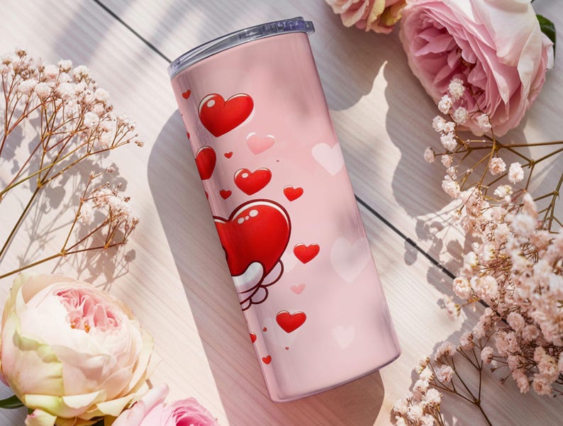 Op de afbeelding: Een roze ge&iuml;soleerde beker versierd met rode en roze harten. De beker heeft een zilveren deksel en is omgeven door roze rozen en witte bloemen, wat een romantisch thema cre&euml;ert. De beker is ontworpen voor dranken.